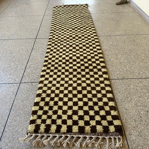 May include: A long, rectangular rug with a checkered pattern in brown and cream. The rug has a fringe border.
