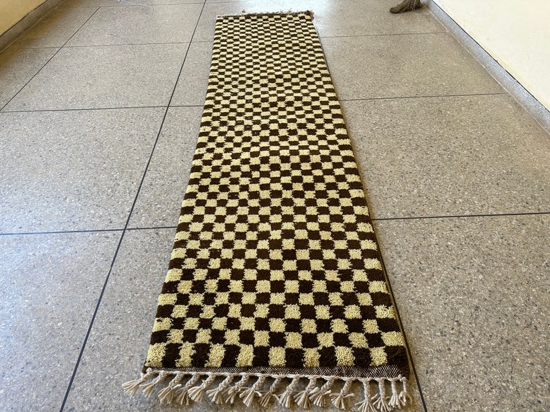 May include: A long, rectangular rug with a checkered pattern in brown and cream. The rug has a fringe border.