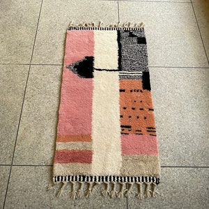 May include: A pink, white, and black geometric patterned rug with fringe on the ends. The rug has a modern design and is made of wool.