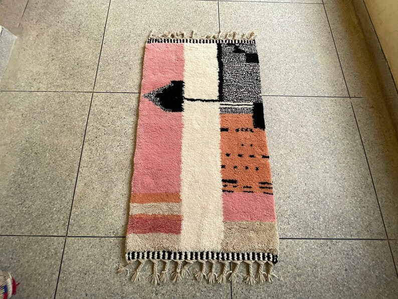 May include: A pink, white, and black geometric patterned rug with fringe on the ends. The rug has a modern design and is made of wool.