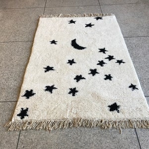 May include: A white rug with a black crescent moon and stars design. The rug has a fringe border.