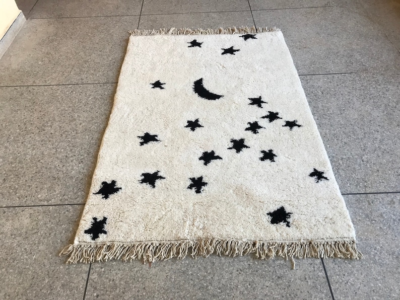 May include: A white rug with a black crescent moon and stars design. The rug has a fringe border.