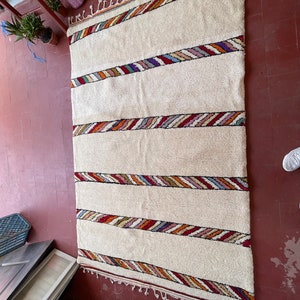 Puede incluir: Una alfombra blanca con rayas de colores tejidas en el dise&ntilde;o. La alfombra tiene un borde de flecos.