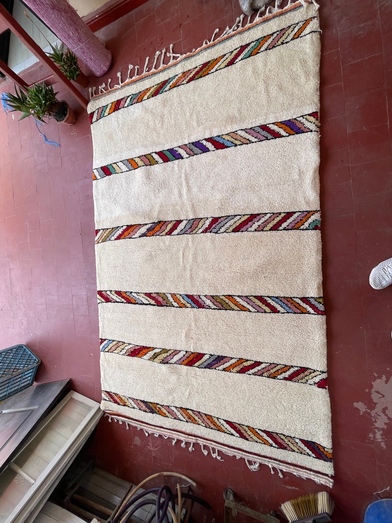 Puede incluir: Una alfombra blanca con rayas de colores tejidas en el dise&ntilde;o. La alfombra tiene un borde de flecos.
