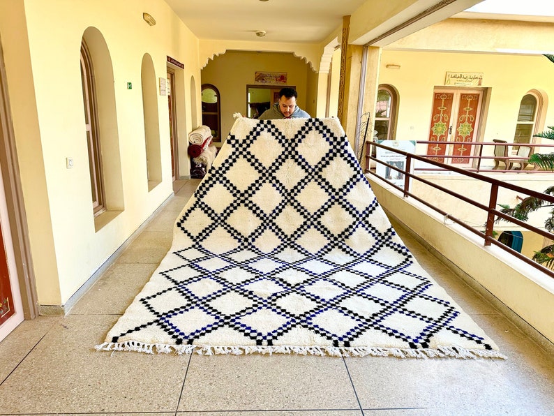 May include: A white and black Moroccan rug with a diamond pattern. The rug has a fringe border and is being held up by a person.
