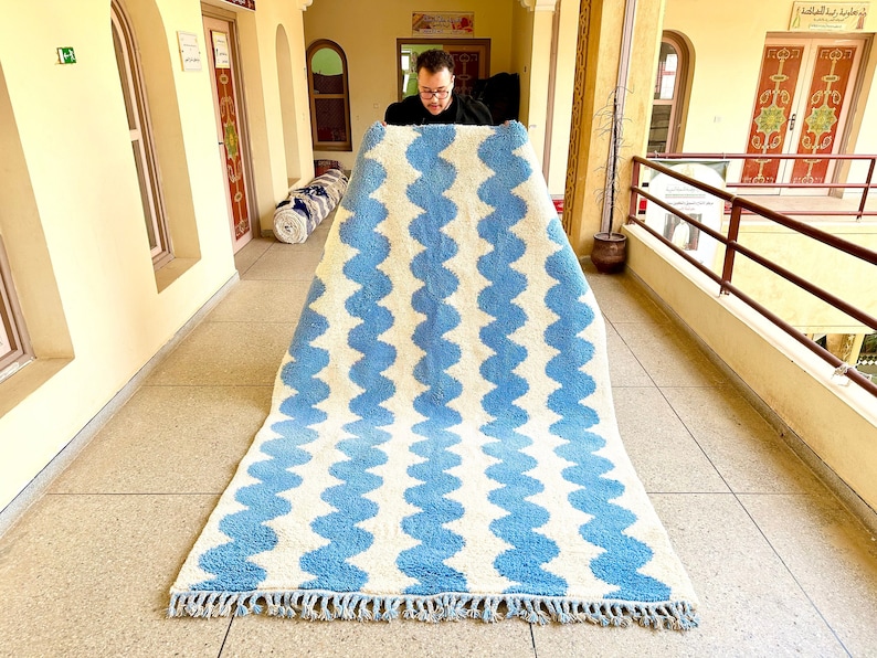 Pu&ograve; includere: Un grande tappeto rettangolare con un motivo a onde ripetute in azzurro e bianco sporco. Il tappeto ha un bordo con frange. Il design &egrave; una dichiarazione grafica audace per qualsiasi stanza.