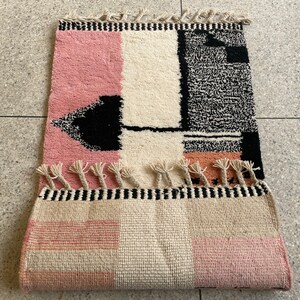 May include: A pink, white, and black geometric patterned rug with fringe. The rug has a black and white geometric design in the center and a pink and white checkered pattern on the bottom.