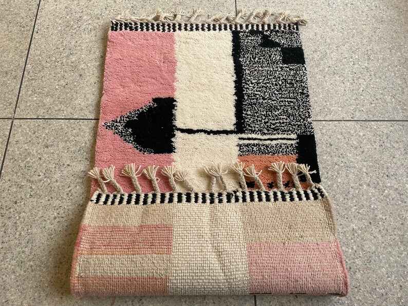 May include: A pink, white, and black geometric patterned rug with fringe. The rug has a black and white geometric design in the center and a pink and white checkered pattern on the bottom.