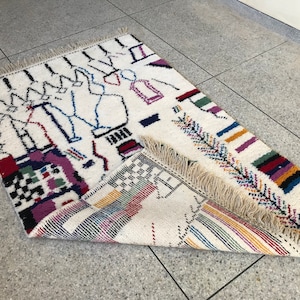 May include: A white rug with a colorful geometric pattern. The rug has a fringe border and is made of wool.