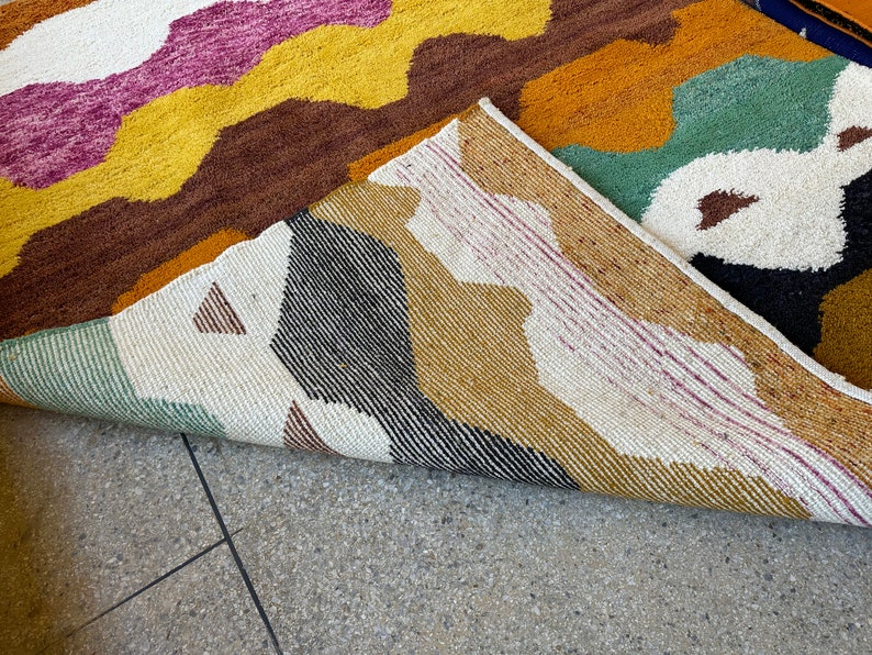 May include: A close-up of a colorful rug with a geometric pattern. The rug is folded in half, revealing the underside of the rug. The rug is made of a soft, woven material.