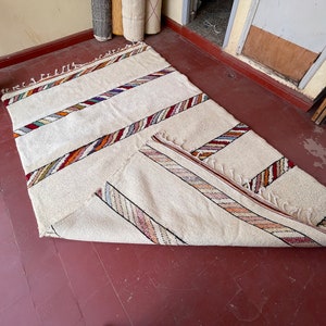 Puede incluir: Una alfombra blanca con rayas de colores tejidas en la tela. La alfombra est&aacute; doblada por la mitad y colocada sobre un suelo de madera.
