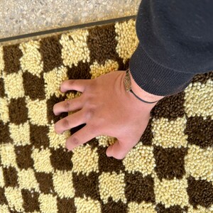 May include: A brown and cream checkered rug with a hand resting on the surface.