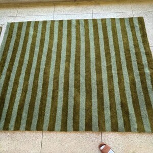May include: A large rectangular rug with vertical stripes in shades of olive green and light green. The rug is placed on a tiled floor, with a smaller patterned rug visible on the left.