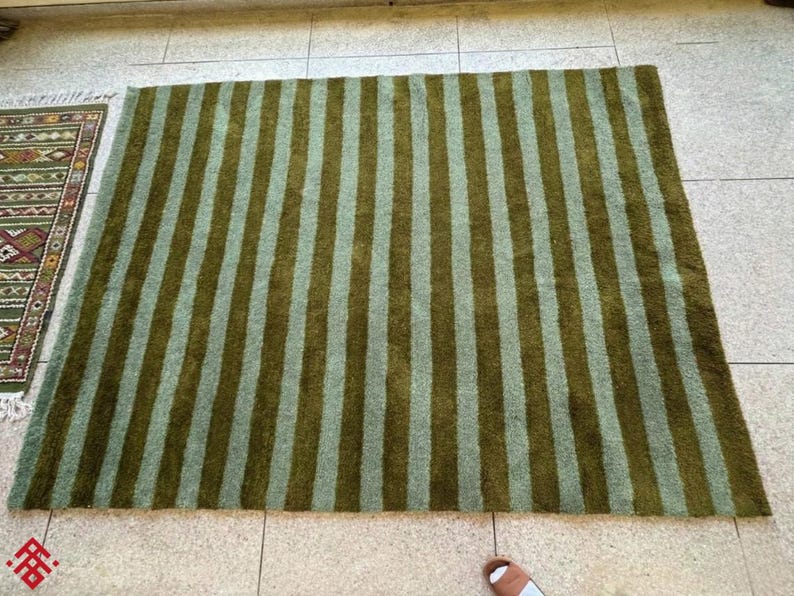 May include: A large rectangular rug with vertical stripes in shades of olive green and light green. The rug is placed on a tiled floor, with a smaller patterned rug visible on the left.