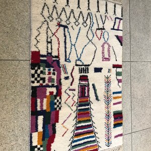 May include: A white rug with a colorful geometric design. The rug has a fringe border and features black, red, blue, green, yellow, and purple colors.