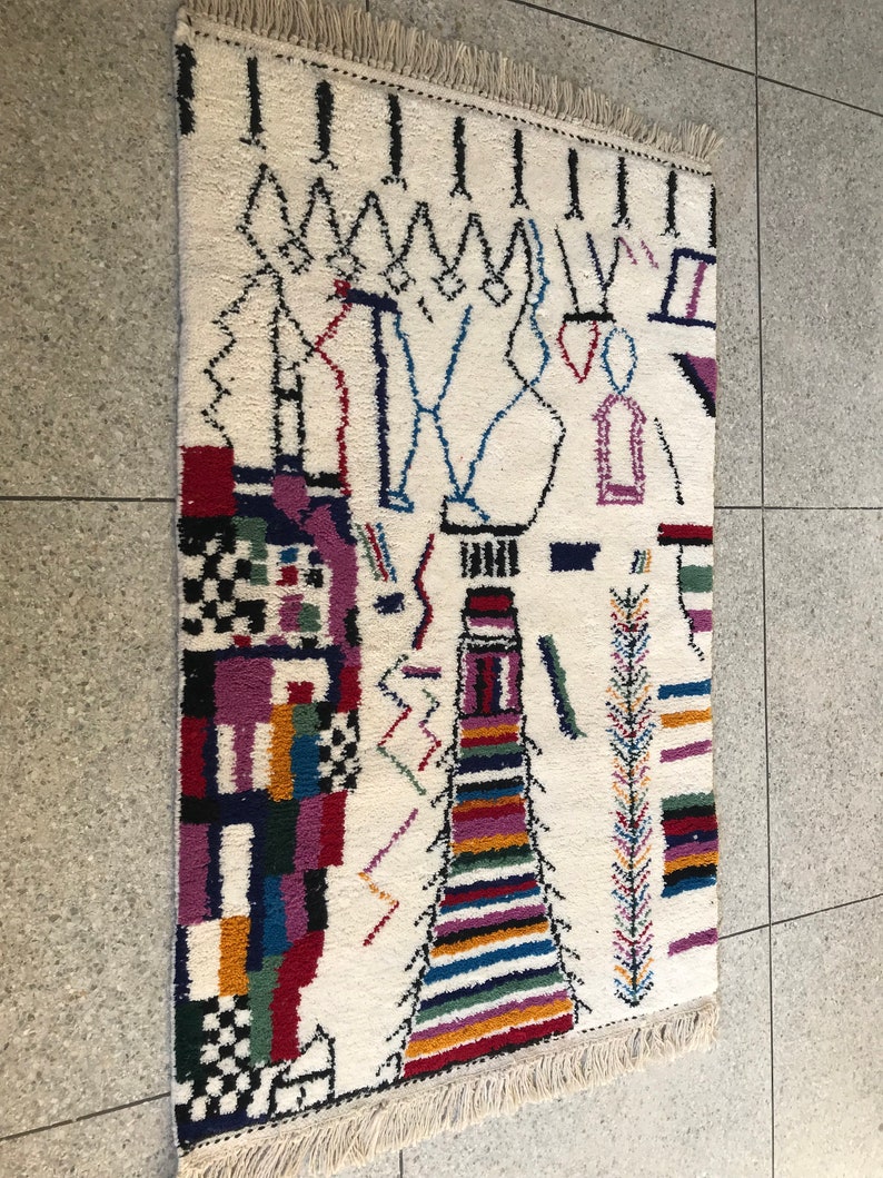 May include: A white rug with a colorful geometric design. The rug has a fringe border and features black, red, blue, green, yellow, and purple colors.