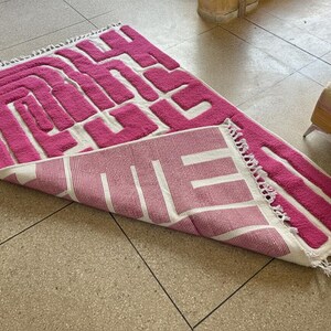 May include: A pink and white geometric patterned rug with a textured design. The rug is rectangular with a fringe on one edge. The design features a maze-like pattern in pink against a white background.