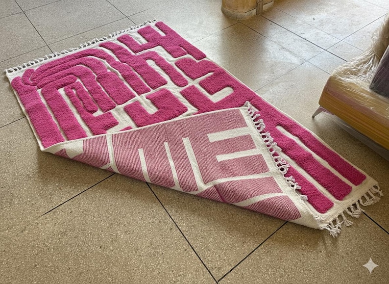 May include: A pink and white geometric patterned rug with a textured design. The rug is rectangular with a fringe on one edge. The design features a maze-like pattern in pink against a white background.