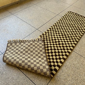 May include: A brown and cream checkered rug with a fringe edge. The rug is folded in half and lying on a tiled floor.