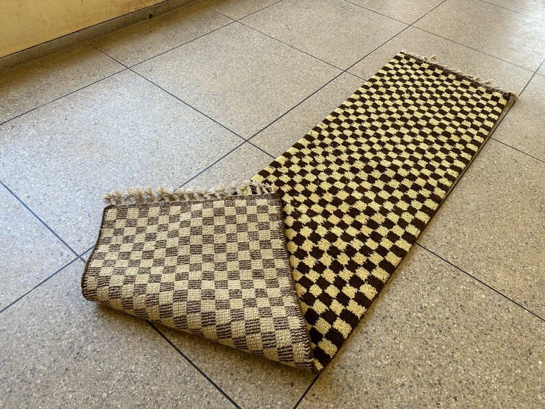 May include: A brown and cream checkered rug with a fringe edge. The rug is folded in half and lying on a tiled floor.