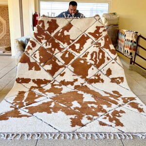 May include: A white and brown Moroccan rug with a geometric pattern. The rug has a fringe border and is made of wool.