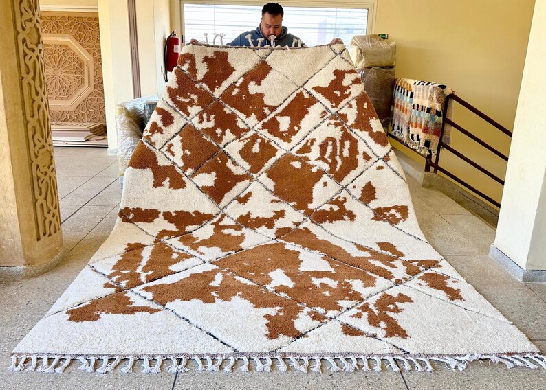 May include: A white and brown Moroccan rug with a geometric pattern. The rug has a fringe border and is made of wool.