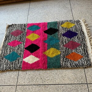 K&ouml;nnte beinhalten: Ein bunter Teppich mit einem geometrischen Muster aus Rauten in verschiedenen Farben, darunter Pink, Schwarz, Gelb, T&uuml;rkis und Orange. Der Teppich hat einen wei&szlig;en Fransenrand.