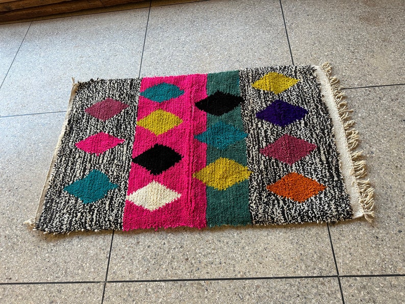 K&ouml;nnte beinhalten: Ein bunter Teppich mit einem geometrischen Muster aus Rauten in verschiedenen Farben, darunter Pink, Schwarz, Gelb, T&uuml;rkis und Orange. Der Teppich hat einen wei&szlig;en Fransenrand.