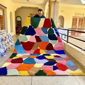 Puede incluir: Una alfombra abstracta colorida con un diseño de patchwork que presenta varias formas y colores, incluyendo rojo, azul, verde, amarillo, naranja y rosa. La alfombra está siendo sostenida por una persona en un pasillo.