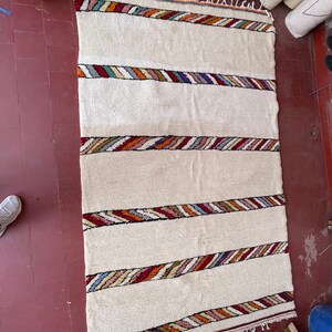 Puede incluir: Una alfombra blanca tejida con rayas de colores. La alfombra tiene un borde de flecos y est&aacute; hecha de fibras naturales.