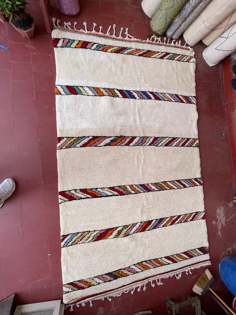 Puede incluir: Una alfombra blanca tejida con rayas de colores. La alfombra tiene un borde de flecos y est&aacute; hecha de fibras naturales.
