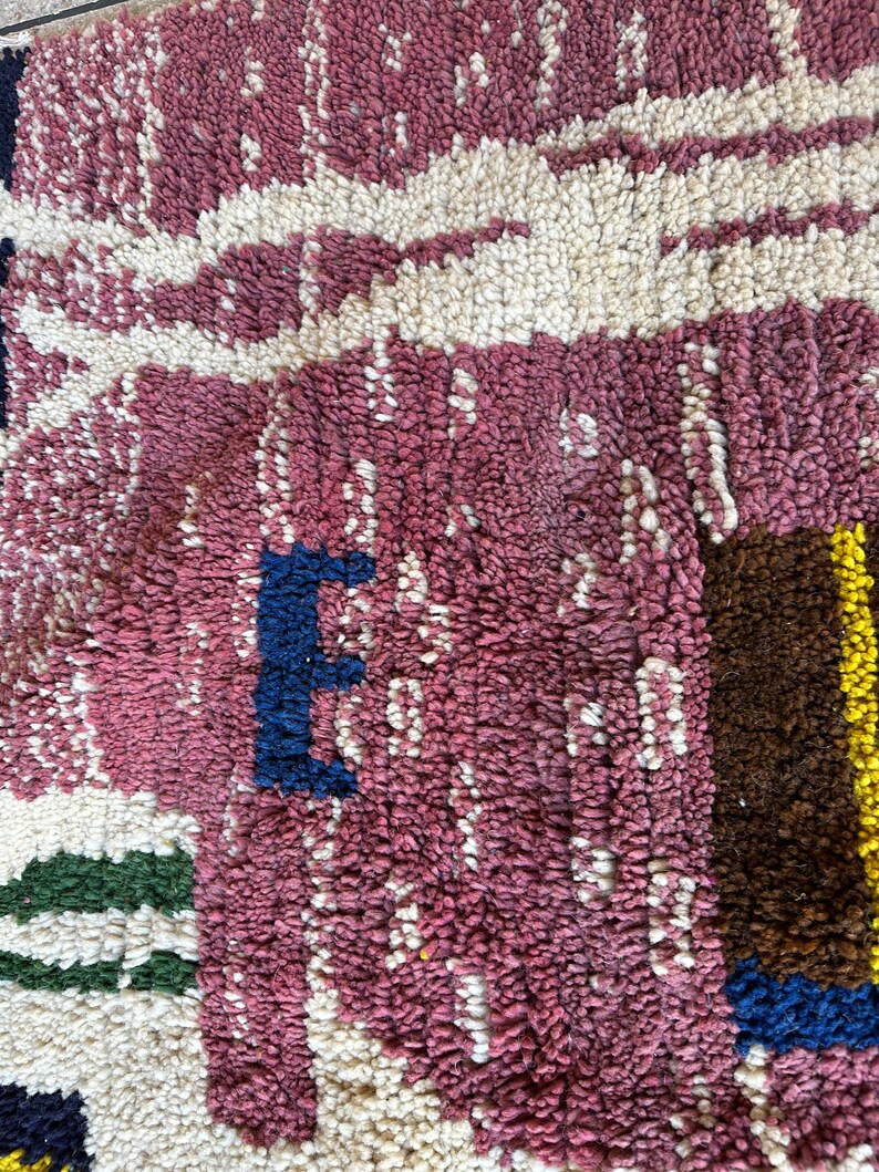 Puede incluir: Primer plano de una alfombra tejida de color rosa, blanco, azul, verde, marr&oacute;n y amarillo con un patr&oacute;n geom&eacute;trico. La alfombra est&aacute; hecha de hilo grueso y texturizado.