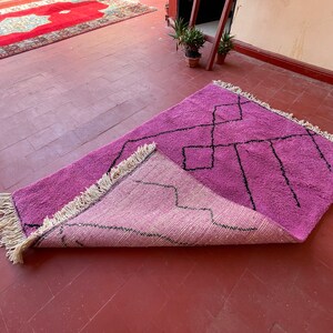 May include: A pink and light pink handwoven rug with a geometric pattern in black. The rug has fringe on all sides and is folded in half.
