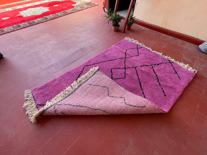 May include: A pink and light pink handwoven rug with a geometric pattern in black. The rug has fringe on all sides and is folded in half.