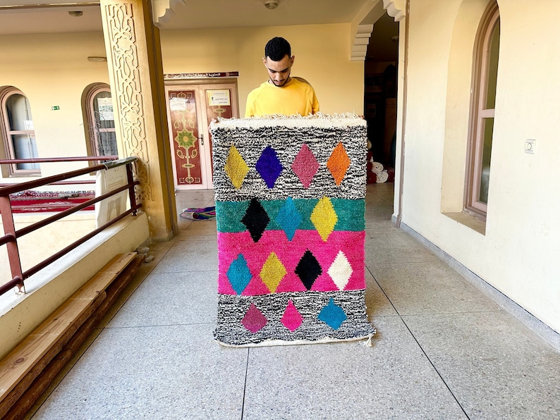 K&ouml;nnte beinhalten: Ein bunter marokkanischer Teppich mit einem geometrischen Diamantenmuster in den Farben Rosa, Blau, Gelb, Gr&uuml;n und Schwarz. Der Teppich wird von einer Person gehalten.