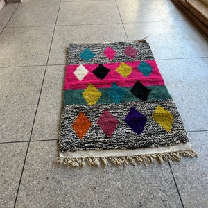 K&ouml;nnte beinhalten: Ein bunter Teppich mit einem geometrischen Muster aus Rauten in den Farben Pink, Blau, Gelb, Orange, Lila und Schwarz. Der Teppich hat einen wei&szlig;en Fransenrand.