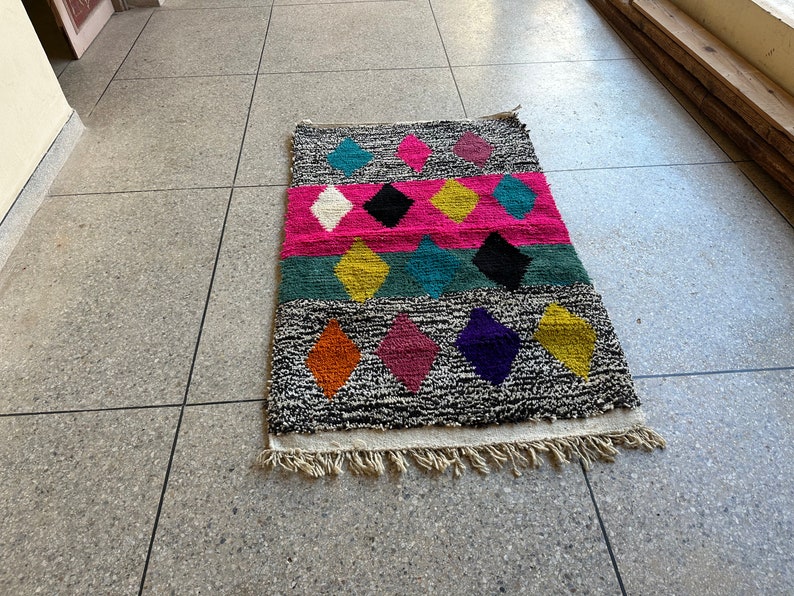 K&ouml;nnte beinhalten: Ein bunter Teppich mit einem geometrischen Muster aus Rauten in den Farben Pink, Blau, Gelb, Orange, Lila und Schwarz. Der Teppich hat einen wei&szlig;en Fransenrand.