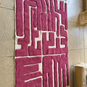 May include: A rectangular rug with a geometric design in shades of fuchsia and white. The rug features a textured, raised pattern in fuchsia against a white background. The edges are finished with a fringe.