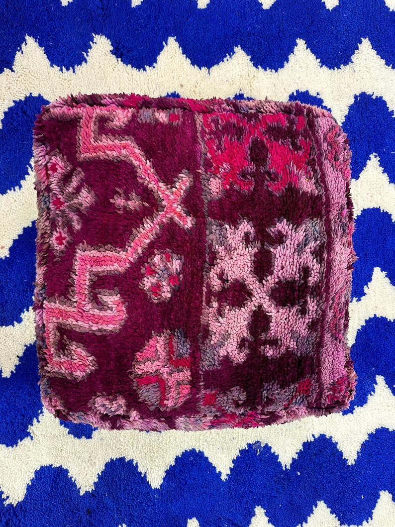Pu&ograve; includere: Un pouf quadrato e morbido nei toni del magenta scuro, fucsia e rosa chiaro. La superficie strutturata presenta motivi geometrici e floreali. Il pouf &egrave; su un tappeto a zig-zag blu e bianco.