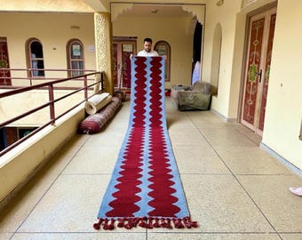 May include: A long, rectangular runner rug featuring a repeating vertical wavy pattern in deep red against a light blue background. The rug is finished with short, thick red tassels along the bottom edge, creating a textured and symmetrical design.