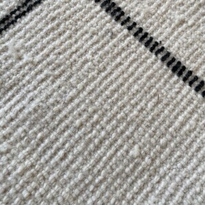 May include: Close-up of a white woven rug with black stripes. The rug has a textured surface and a subtle sheen.