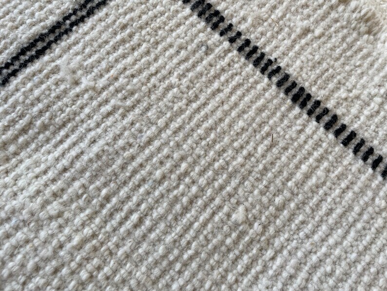 May include: Close-up of a white woven rug with black stripes. The rug has a textured surface and a subtle sheen.