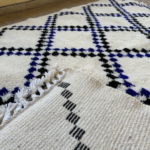 May include: White rug with a geometric pattern of black and blue squares. The rug has a fringe border.