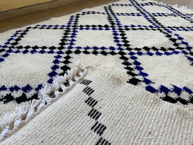 May include: White rug with a geometric pattern of black and blue squares. The rug has a fringe border.