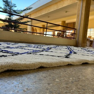 May include: A white rug with a black and blue geometric pattern. The rug is lying on a light brown tiled floor.