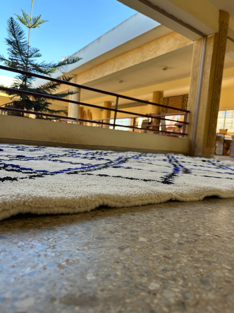 May include: A white rug with a black and blue geometric pattern. The rug is lying on a light brown tiled floor.