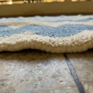 Pu&ograve; includere: Primo piano di un tappeto morbido e strutturato con un motivo a spina di pesce. Il tappeto presenta fasce alternate di azzurro e bianco sporco, creando un design a forma di onda. Il bordo del tappeto &egrave; visibile, mostrando lo spessore e la consistenza soffice.