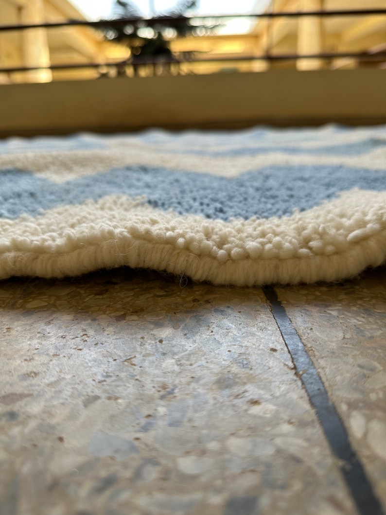 Pu&ograve; includere: Primo piano di un tappeto morbido e strutturato con un motivo a spina di pesce. Il tappeto presenta fasce alternate di azzurro e bianco sporco, creando un design a forma di onda. Il bordo del tappeto &egrave; visibile, mostrando lo spessore e la consistenza soffice.