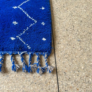 Puede incluir: Una alfombra azul con un patr&oacute;n geom&eacute;trico blanco y flecos. La alfombra est&aacute; sobre una superficie texturizada gris claro.