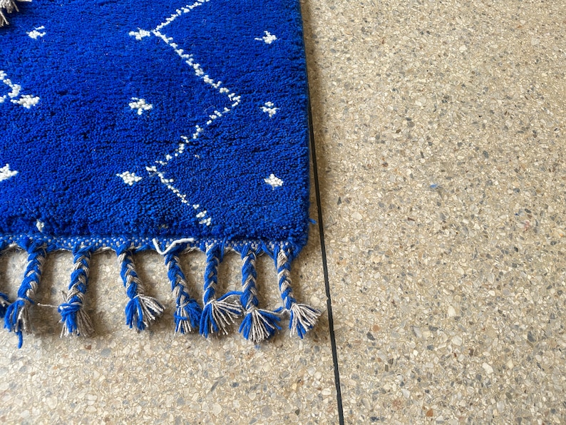 Puede incluir: Una alfombra azul con un patr&oacute;n geom&eacute;trico blanco y flecos. La alfombra est&aacute; sobre una superficie texturizada gris claro.